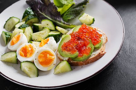 Téléchargez les photos : Breakfast. Healthy open sandwich on  toast with avocado and red caviar, boiled eggs, cucumber salad on white plate. Healthy protein food. - en image libre de droit