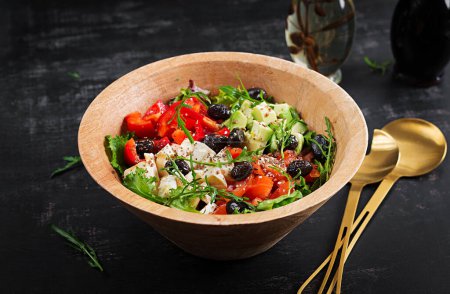 Foto de Fresh salad with avocado, tomatoes, mozzarella cheese, sweet peppers and black olives on wooden bowl. - Imagen libre de derechos