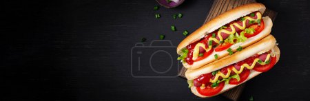 Téléchargez les photos : Hot dog with grilled sausage, tomato and lettuce on dark background. American hotdog. Top view, banner - en image libre de droit