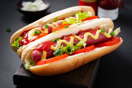 Foto de Hot dog with grilled sausage, tomato and lettuce on dark background. American hotdog - Imagen libre de derechos