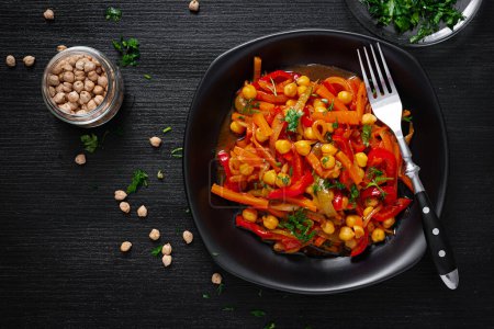 Photo for Vegetable stew of chickpeas, carrot, sweet peppers, onions, garlic and tomato sauce with lemon on the dark table. Oriental cuisine. Top view - Royalty Free Image
