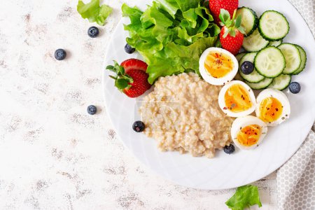 Photo for Breakfast oatmeal porridge with boiled eggs, cucumber and strawberry. Healthy balanced food. Top view, flat lay - Royalty Free Image