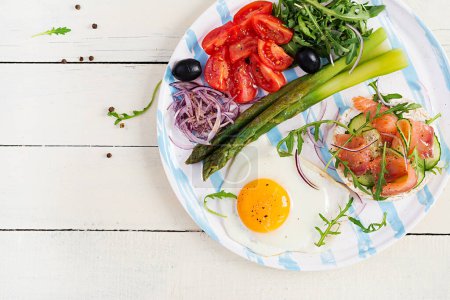 Photo for Ketogenic breakfast. Breakfast. Fried egg, crisp loaves with salmon, green asparagus, fresh tomatoes and red onions on white plate. Top view, flat lay - Royalty Free Image