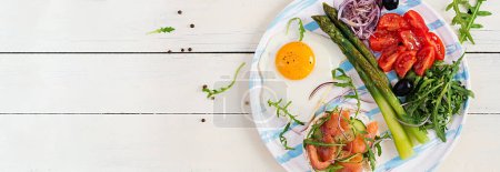 Photo for Ketogenic breakfast. Breakfast. Fried egg, crisp loaves with salmon, green asparagus, fresh tomatoes and red onions on white plate. Top view, banner - Royalty Free Image
