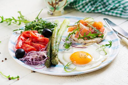 Photo for Ketogenic breakfast. Breakfast. Fried egg, crisp loaves with salmon, green asparagus, fresh tomatoes and red onions on white plate. - Royalty Free Image