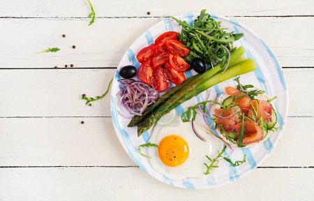 Photo for Ketogenic breakfast. Breakfast. Fried egg, crisp loaves with salmon, green asparagus, fresh tomatoes and red onions on white plate. Top view, flat lay - Royalty Free Image