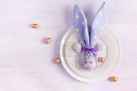 Photo for Easter table setting. White plate with a napkin folded in the shape of a rabbit, Easter and chocolate eggs on a pink background. Happy Easter holiday concept. Top view, flat lay - Royalty Free Image