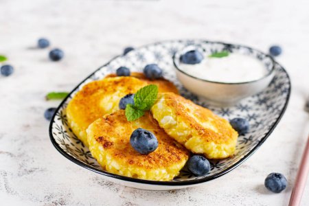 Téléchargez les photos : Crêpes au fromage cottage avec crème sure et bleuets sur un fond clair. Petit déjeuner ou déjeuner. - en image libre de droit