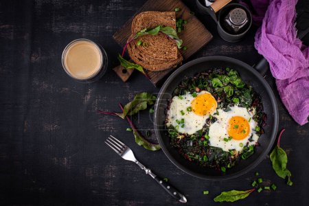 Foto de El desayuno. Huevos fritos con hojas de remolacha verde, ajo, pimienta y cebolla verde en sartén. Vista superior, plano - Imagen libre de derechos