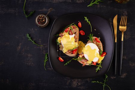 Foto de El desayuno. Sandwich con jamón, huevo escalfado, cebolla y rúcula en un plato sobre fondo oscuro. Vista superior - Imagen libre de derechos