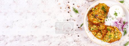 Foto de Buñuelos de calabacín con cebolla roja, ajo y hierbas. Panqueques vegetarianos de calabacín y crema agria en la mesa blanca. Vista superior, banner - Imagen libre de derechos