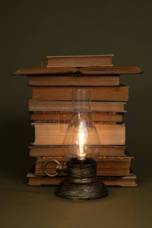 Foto de Libros antiguos y una lámpara con una luz ardiente sobre un fondo de olivo. Lámpara estilizada contemporánea. - Imagen libre de derechos