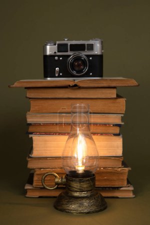 Foto de Libros antiguos y una lámpara de mano con bombilla, estilizada como antigua. Hay una vieja cámara encima de los libros. Objetos colocados sobre un fondo de olivo. - Imagen libre de derechos