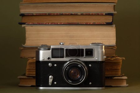 Foto de Una vieja cámara analógica y viejos libros amarillentos apilados detrás de la cámara. Artículos sobre fondo verde oliva. - Imagen libre de derechos