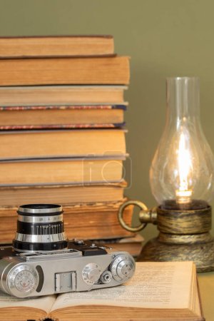 Foto de Un viejo libro abierto en el que se encuentra una cámara de fotos analógica. En el fondo, una pila de libros antiguos y una lámpara estilizada como una lámpara de queroseno antiguo. Artículos colocados sobre un fondo verde oliva. - Imagen libre de derechos