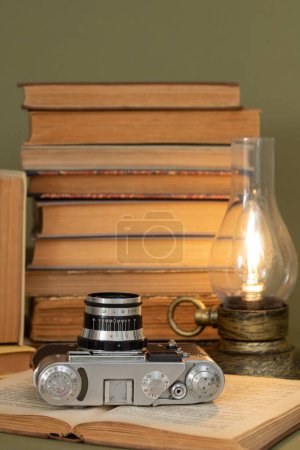 Foto de Un viejo libro abierto en el que se encuentra una cámara de fotos analógica. En el fondo, una pila de libros antiguos y una lámpara estilizada como una lámpara de queroseno antiguo. Artículos colocados sobre un fondo verde oliva. - Imagen libre de derechos