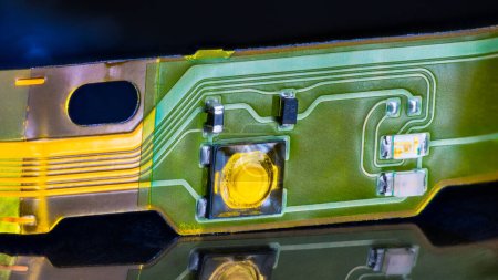 Nahaufnahme einer elektronischen Flexschaltung mit Reflexion auf schwarzem Hintergrund. Flache Kunststoffleiste mit kleinen Aufputzkomponenten auf grün-gelber flexibler Leiterplatte aus demontierten Kopfhörern.