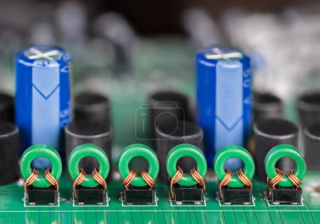 Foto de Primer plano de bobinas de núcleo toroidal y condensadores electrolíticos azules en una PCB verde. Fila de inductores de transformadores en placa de circuito impreso para aislamiento galvánico de señal de audio o vídeo con fondo borroso. - Imagen libre de derechos