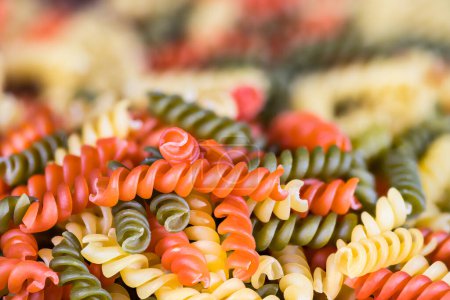 Photo for Closeup of colored spiral twisted fusilli pasta on pile with blurred background. Side view a heap of dry uncooked yellow, red and green eggless rotini of Italian cuisine with tomato or spinach flavor. - Royalty Free Image