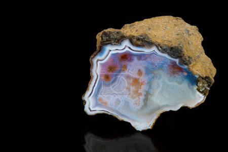 Polished cut of natural agate precious stone with raw brown surface on a black background. Closeup of beautiful smooth icy blue cross section by chalcedony mineral rock with red stains and white border.