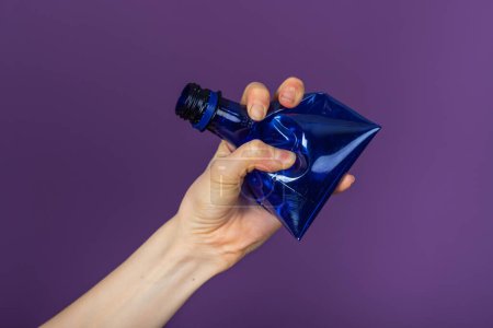 Photo for Womans hand holds a crumpled blue plastic bottle on a purple background - Royalty Free Image