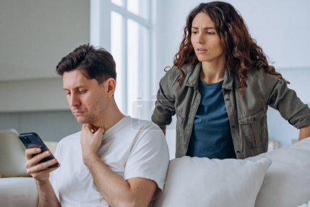 Photo for Young woman looks out over husband shoulder at phone screen and starts quarreling accusing man in cheating in living room - Royalty Free Image