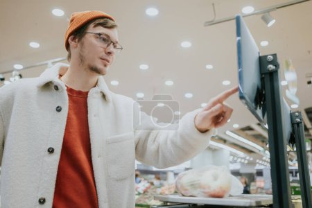 Foto de Un hombre pesa manzanas en una bolsa en un supermercado en una balanza electrónica. Selecciona el producto deseado con el dedo en la pantalla táctil para determinar el peso y el precio - Imagen libre de derechos