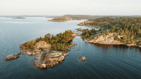 The golden hour sunlight bathes rugged coastal cliffs and evergreen trees, highlighting the intricate textures of the landscape. A serene channel of water weaves through the islands, reflecting the