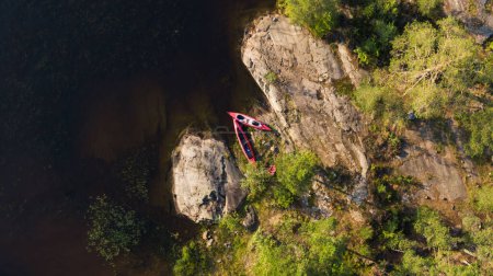 Aerial view reveals a vibrant red kayak resting on the sun-kissed riverbank, nestled among rocks and greenery. The scene captures the essence of outdoor adventure and the tranquility of nature