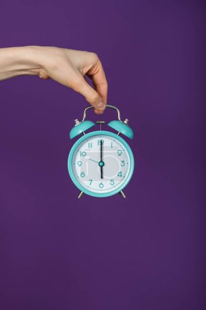 A hand suspends a classic blue alarm clock against a vivid purple background, illustrating the concept of time management. The simple composition emphasizes the importance of punctuality. Hand Holding