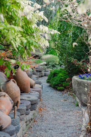  A serene gravel path meanders through a lush garden, flanked by antique pottery and vibrant plants. It invites a peaceful stroll amidst the greenery.