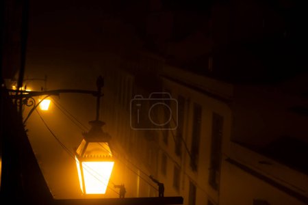 Mysterious Foggy Night in Old Town with Vintage Street Lamp. A warm glow emanates from an antique street lamp on a foggy night, the mist veiling the outlines of buildings behind.