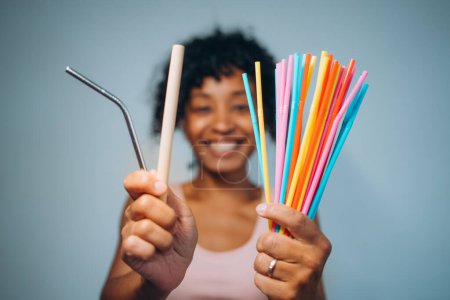 Eine fröhliche asiatisch-afrikanische Frau vergleicht einen wiederverwendbaren Metallstrohhalm mit bunten Plastikstrohhalmen und betont damit die Wahl zwischen nachhaltigen und Einwegprodukten.