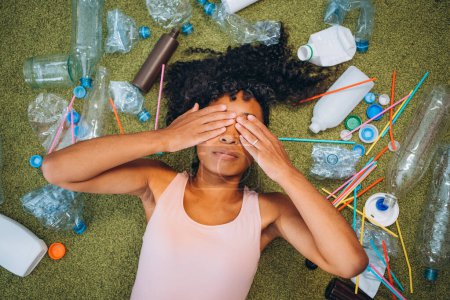 Eine Frau liegt auf dem Teppich, umgeben von einer Reihe weggeworfener Plastikflaschen und -riemen, und verdeckt entsetzt ihre Augen. Von Plastikmüll überwältigt