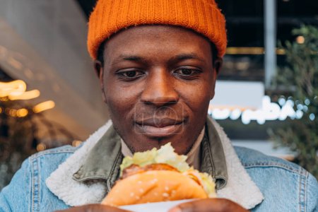 African American Man in Beanie Holding and Looking at a Delicious Burger with Anticipation. A close-up of a man with a warm smile, ready to enjoy a juicy burger, captured in a vibrant urban eatery