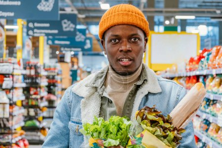 Young African American Man Shopping for Fresh Produce in Grocery Store. A man in a trendy orange beanie and denim jacket holds fresh bread and leafy greens while shopping in a grocery store