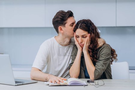 In a touching display of support, a man comforts his partner who appears concerned while managing finances at home, highlighting the emotional aspect of financial planning