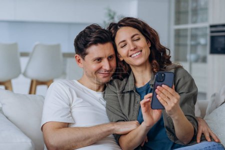 Smiling Couple Taking a Selfie on Couch in a Chic Living Room. A smiling couple enjoys a cozy moment on the couch, looking at a smartphone together