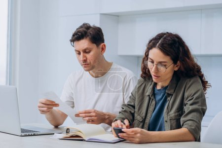 A focused couple sits at a table with a laptop and documents, engaged in managing their household finances, embodying teamwork in everyday life. The woman writes down all expenses in a notebook