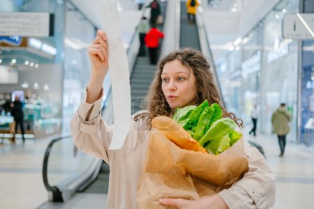 A woman looks surprised as she examines a long grocery receipt, while holding a paper bag of fresh produce, in a shopping mall.