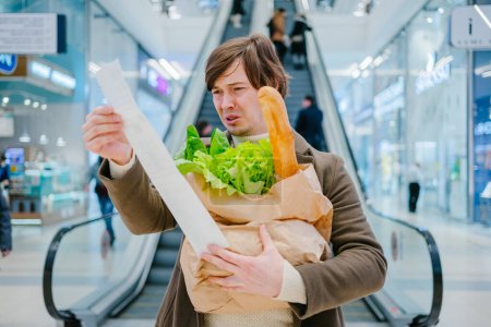 A man in a casual outfit is visibly perplexed while looking at a lengthy grocery receipt, holding a bag of fresh food in a shopping center.