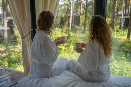 Two woman in white robes enjoy a peaceful morning, sipping coffee while seated on a bed, gazing out to a lush forest from a cozy cabin window. Friends are enjoying their vacation in a country house