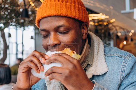 Afroamerikaner in orangefarbener Mütze genießen den ersten Bissen eines saftigen Burgers. Ein zufriedener Mann nimmt einen großen Bissen von einem Burger und genießt das Essen im gemütlichen Ambiente eines städtischen Lokals
