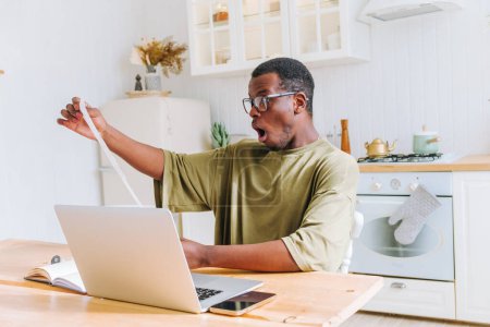 Schockierter Afroamerikaner mit langer Quittung bei der Arbeit am Laptop. Ein erstaunter Mann mit Brille hält eine übermäßig lange Quittung in der Hand, sein Mund zittert vor Überraschung, als er an einem Küchentisch sitzt.