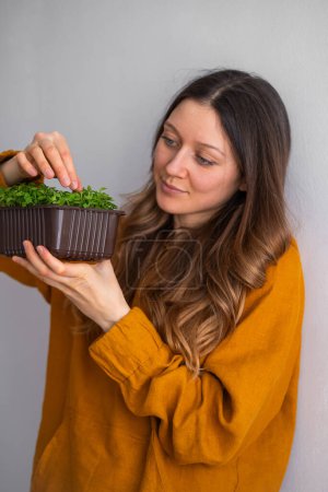 Photo for Woman in a mustard blouse admires a lush pot of microgreens, reflecting a moment of home gardening satisfaction. Arugula cultivation at home, fresh healthy greens rich in vitamins. - Royalty Free Image