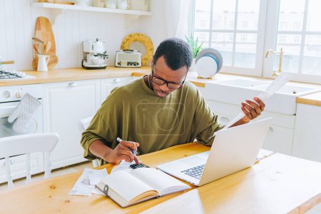 Ein fokussierter afroamerikanischer Mann verwaltet seine Finanzen am Küchentisch mit Laptop und Taschenrechner, mit Quittung in der Hand, was die persönliche Buchhaltung zu Hause veranschaulicht.
