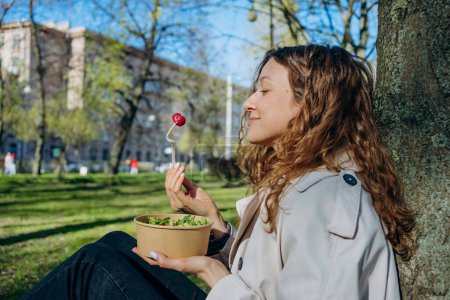 Una joven disfruta de una ensalada fresca y frambuesa al aire libre, relajándose contra un árbol en un soleado parque urbano. 