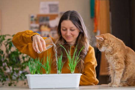 Una mujer sonriente en un suéter de mostaza riega brotes de cebolla verde en una maceta mientras un curioso gato jengibre observa, un momento de felicidad de jardinería en casa