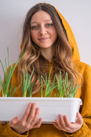 A young woman with blue eyes and a warm smile, wearing a goldenrod hoodie, holds a white planter box of fresh green sprouts. Growing greenery at home by the window, home gardening.