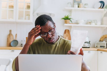 Besorgter Afroamerikaner überprüft Finanzen mit Laptop und Quittung zu Hause. In einer hellen Wohnküche zeigt ein Mann Anzeichen von Stress, als er Rechnungen auf seinem Laptop begutachtet.
